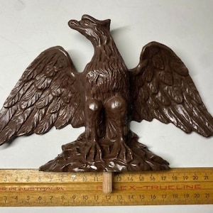 May include: A brown eagle wall plaque with spread wings, perched on a textured base. A wooden ruler is positioned underneath, showing the eagle spans approximately 30.5 cm in width from the tip of one wing to the tip of the other wing.