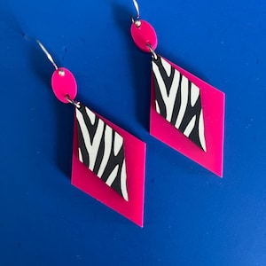 May include: A pair of dangle earrings featuring a hot pink oval top piece connected to a larger hot pink diamond-shaped pendant. A smaller white diamond-shaped piece with a black zebra stripe pattern is layered on top of the larger pink diamond.