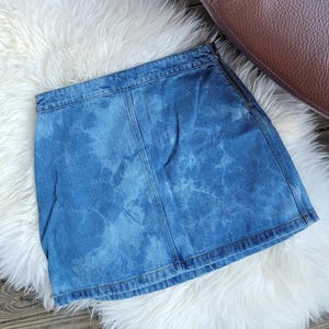 May include: A blue denim mini skirt with a tie-dye effect. The skirt is displayed on a white fluffy rug. The skirt has a classic five-pocket design and is made of denim.