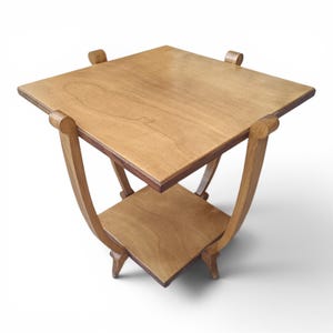 Puede incluir: Una mesa auxiliar cuadrada de madera con un acabado de veta de madera marrón claro en la parte superior y el estante inferior. La mesa está sostenida por cuatro patas curvas que se extienden hacia arriba para unirse a las esquinas superiores.