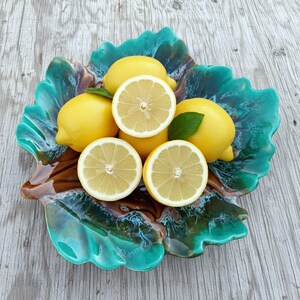 Può includere: Un piatto in ceramica a forma di foglia con una smaltatura lucida color turchese, verde e marrone contenente diversi limoni gialli interi e tagliati a metà. Il piatto presenta bordi organici ondulati e una superficie strutturata.