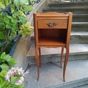 Peut inclure: Une table d'appoint haute en bois brun clair dotée d'un tiroir unique avec une poignée en métal foncé ornée. La table présente un plateau plat aux bords surélevés, une étagère de rangement ouverte sous le tiroir et quatre pieds fins et incurvés.