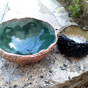 Może przedstawiać: Dwie ręcznie wykonane miski ceramiczne o nieregularnych, teksturowanych powierzchniach zewnętrznych. Większa miska ma błyszczącą, głęboko zieloną glazurę wewnątrz, podczas gdy mniejsza ma ciemną, teksturowaną czarną powierzchnię zewnętrzną i jasne wnętrze.