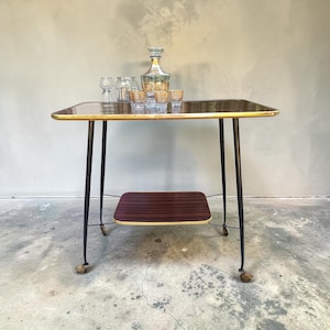 Peut inclure: Un chariot de service à deux niveaux avec un plateau supérieur en bois foncé et une étagère inférieure plus petite, soutenu par quatre pieds fins en métal noir sur roulettes. Une carafe en verre et quatre verres assortis à bandes dorées.