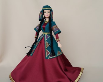 Puede incluir: Una muñeca con un vestido tradicional bordado en verde y dorado con una falda color burdeos y un pañuelo negro en la cabeza.