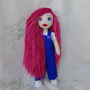 Puede incluir: Muñeca hecha a mano con pelo largo y rosa vibrante y ojos azules. La muñeca lleva una camisa blanca con estampado floral debajo de un mono azul y zapatos blancos. La cara de la muñeca está pintada a mano con labios rojos.