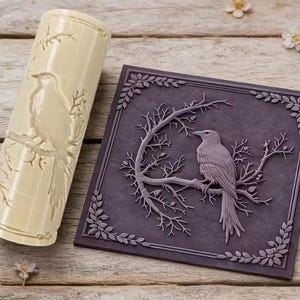 May include: A cylindrical cream-colored rolling pin with an engraved crow and branch pattern, positioned next to a square dark purple tile featuring a raised relief design of a crow perched on a bare tree branch surrounded by a leafy border.
