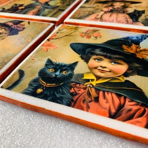 May include: Set of decorative square coasters featuring vintage illustrations of children in witch costumes with black cats. The coasters have a white surface with an orange border. The images have a vintage, antique aesthetic.