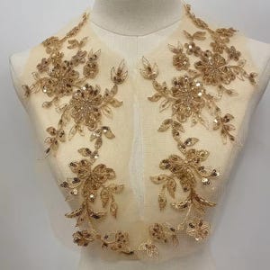 May include: A pair of symmetrical gold floral appliques featuring intricate embroidery, metallic thread, and shimmering sequins on a sheer nude mesh base. The design consists of detailed flower blossoms and leafy vine patterns arranged vertically.