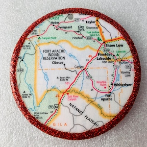 May include: A round ornament featuring a map of the Fort Apache Indian Reservation and surrounding areas. The map is in shades of green, yellow, and white, with a red glitter border. The ornament includes place names and road numbers.