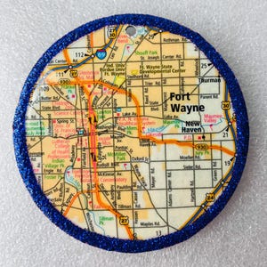 May include: Circular ornament featuring a map of Fort Wayne, Indiana, with a blue glitter border. The map includes street names, parks, and landmarks. The ornament is a decorative item.