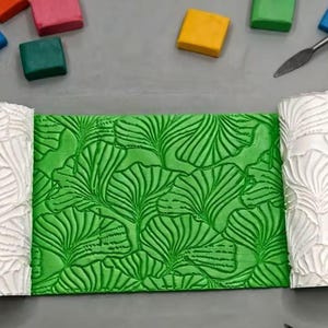 May include: A bright green rectangular slab of clay featuring a deeply embossed ginkgo leaf pattern, flanked on both sides by white cylindrical rollers that display the same intricate leaf design used to texture the clay surface.