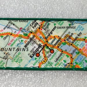 May include: Rectangular magnet featuring a map of the Rocky Mountains, with the text "ROCKY MOUNTAINS" and "ROCKY" visible. The map includes roads, towns, and geographical features, framed by a green border.