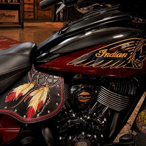 May include: A black and deep red motorcycle fuel tank featuring a gold Indian Motorcycle logo and head graphic. Below the tank, a black leather side panel displays a detailed dreamcatcher design with red and yellow feathers and metal studs.