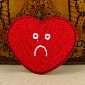 May include: Red felt heart-shaped patch with a white embroidered sad face.