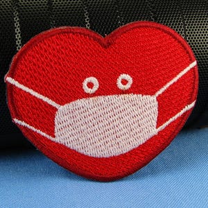 May include: Embroidered red heart patch with a white face mask and two small white eyes. The patch is on a blue surface, with a black speaker in the background. The patch is a symbol of love and health.