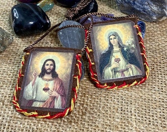 Puede incluir: Dos escapularios marrones rectangulares con imágenes religiosas. Uno representa a Jesús con el sagrado corazón y el otro a la Virgen María sosteniendo lirios. Ambos tienen bordes con costuras rojas y amarillas y cordones marrones.