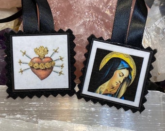 May include: Two square ornaments with black scalloped felt borders and black ribbon loops. One ornament features a red heart pierced by swords and topped with flames. The other ornament displays a portrait of a figure with a yellow halo.