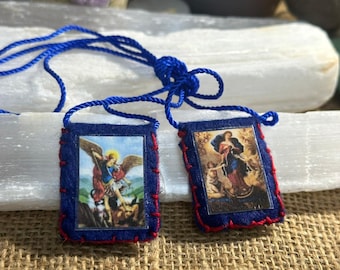 Può includere: Due scapolari rettangolari in feltro blu scuro collegati da un cordino blu. Ogni scapolare presenta un'immagine religiosa cucita con filo rosso. L'immagine a sinistra raffigura un angelo alato, quella a destra una figura in abiti con cherubini.