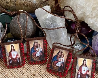 Puede incluir: Cuatro escapularios de tela marrón con bordes cosidos en rojo, cada uno con una imagen religiosa. Dos representan a Jesús con un sagrado corazón y dos representan a la Virgen María sosteniendo al niño Jesús. Cada escapulario cuelga de un cordón marrón.