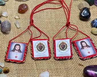 May include: Four square fabric scapulars with red cord strings. Two feature an image of Jesus with a Sacred Heart, and two feature the Sacred Heart emblem with Spanish text reading Detente el Corazón de Jesús está conmigo.