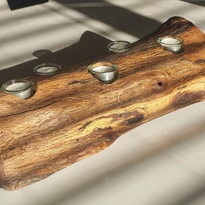 May include: A rustic wooden candle holder with five glass votive candle holders. The wood has a natural, warm brown tone with visible grain and knots. The candle holder is placed on a white surface, with shadows cast across it.