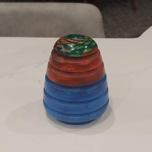 May include: A decorative, layered object with a blue base, red middle, and green top. The object has a rounded shape with horizontal ridges on the blue section. The top section has a textured, scale-like pattern.