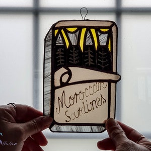 May include: A stained glass window hanging features a brown rectangular frame with a yellow and white design at the top. The text "Moroccan Sardines" is written in black cursive inside the frame.