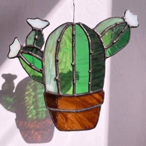 May include: A stained glass cactus suncatcher featuring vibrant green glass segments with dark metal soldering. The cactus is potted in a brown glass base and topped with small white glass flowers, suspended by a thin wire loop.