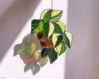 May include: A stained glass suncatcher depicting a potted plant with variegated leaves in shades of lime green and dark forest green. The pot is a solid brown glass, and the entire piece is held together by dark metal solder lines.