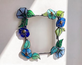 May include: A square stained glass picture frame featuring blue and light blue flowers with green leaves. The frame is constructed with dark metal soldering and includes small amber-colored glass accents in the center of the flowers.