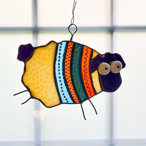 May include: A whimsical stained glass sheep ornament. The sheep features a yellow body with a colorful striped design in teal, orange, and blue. It has large, googly eyes and is suspended by a wire loop.