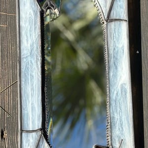 May include: Rectangular stained glass mirror with a spiderweb design. The mirror has a clear, oval-shaped reflective surface. The frame is white and blue with black accents, and features two iridescent butterfly-like shapes.