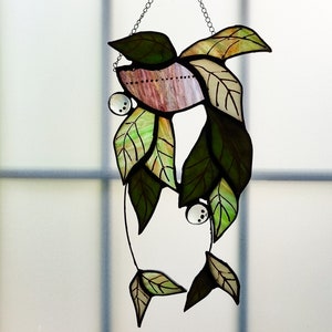 May include: A stained glass window hanging with a design of green and brown leaves with a pink and white center. The leaves are connected by a silver chain.