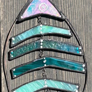 May include: A stained glass fish skeleton suncatcher with a black metal frame. The fish has iridescent teal, green, and blue glass pieces. The top and bottom pieces are triangular, and the middle pieces are horizontal.