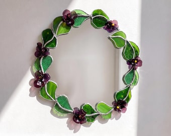 May include: A circular stained glass wreath featuring translucent green leaves and deep purple flowers with small yellow centers. The individual glass pieces are joined together with silver-toned metal solder, creating a continuous floral ring.