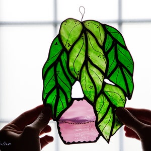 May include: A stained glass window hanging with a pink pot and green leaves. The leaves are outlined in black and have small black dots on them.