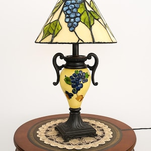 May include: A decorative table lamp with a stained glass shade featuring a blue grape cluster design. The lamp has a yellow base with a grape motif and black accents. It sits on a round, wooden table with a decorative lace doily.