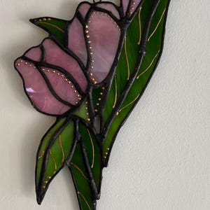 May include: Stained glass floral art piece featuring a pink flower with green leaves. The flower has a delicate, translucent appearance with gold accents. The piece is outlined in black and is designed to hang.