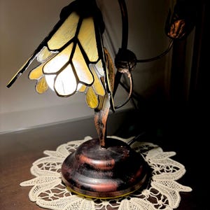 May include: A stained glass table lamp with a floral design. The lamp has a yellow and brown stained glass shade and a brown metal base. The lamp is sitting on a white crocheted doily.