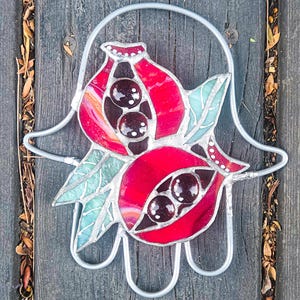 May include: A stained glass Hamsa hand with two red pomegranates. The pomegranates have black seeds and green leaves.
