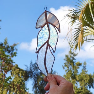 May include: A stained glass feather with iridescent blue and white glass pieces. The feather is held in a hand against a blue sky background.