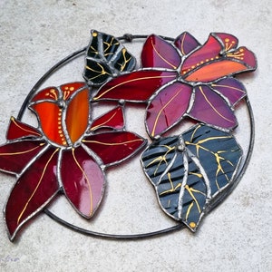 May include: Stained glass floral art piece featuring three flowers in shades of red, orange, and purple, with green and black leaves. The flowers and leaves are outlined in silver and accented with gold details, set within a circular metal frame.