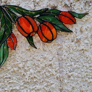 May include: A stained glass art piece depicting a branch with green leaves and orange fruit. The piece is mounted on a white textured wall.