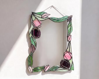 May include: A rectangular stained glass mirror featuring a decorative frame adorned with green leaves and pink and dark purple tulip-like flower buds. The glass pieces are joined by dark metal soldering, and a thin wire is attached for hanging.