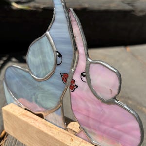 May include: A stained glass sculpture of two elephants, one blue and one pink, facing each other. The elephants have smiling faces and are mounted on a wooden base. The glass has a textured, iridescent quality.
