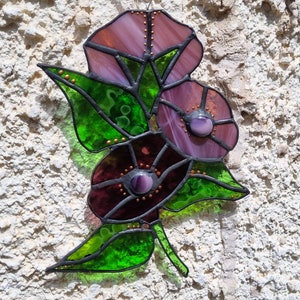 May include: A stained glass window hanging features three pink and purple flowers with green leaves. The flowers have a textured, iridescent look.