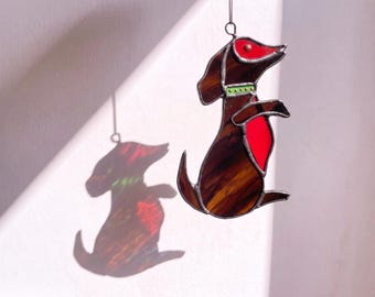 May include: A stained glass suncatcher shaped like a dachshund dog. The dog is brown with a red ear, a red chest patch, and a green collar with small gold dots. It is suspended by a metal ring and casts a colorful shadow on the wall.