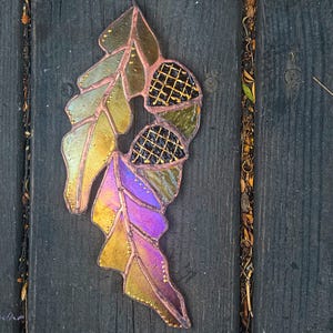 May include: A handcrafted stained glass leaf and acorn pendant. The pendant features a combination of colors, including gold, green, and purple. The acorn caps have a black and gold grid pattern. The pendant is designed to be hung.