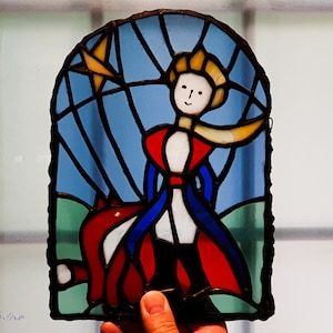 May include: A stained glass window depicting The Little Prince standing on a planet with a star above him. The prince is wearing a red and blue cape and a white shirt. The planet is green and blue.